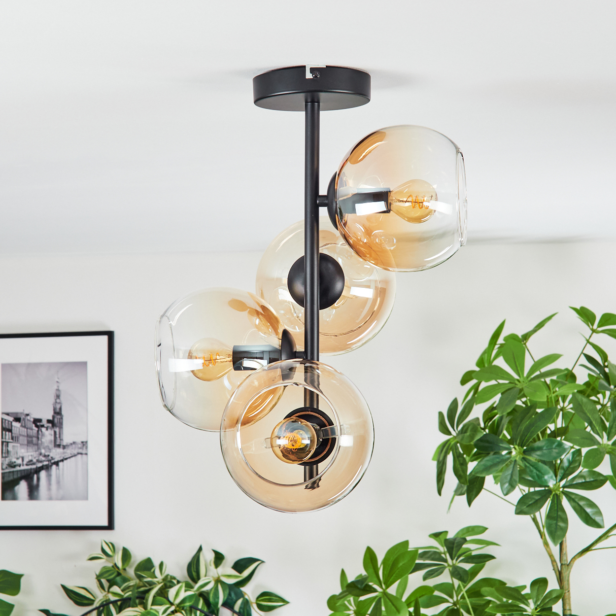 Eine moderne Deckenlampe mit vier bernsteinfarbenen Glaskugeln. Die Lampe ist schwarz, mit einer weißen Decke. Pflanzen und ein gerahmtes Bild sind im Hintergrund.