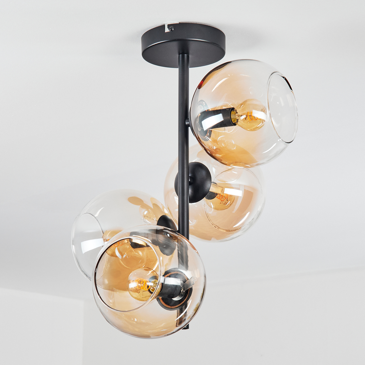 Eine moderne Deckenlampe mit vier bernsteinfarbenen Glaskugeln, in denen sich jeweils eine Glühbirne befindet. Die Leuchte ist schwarz und an einer weißen Decke befestigt.