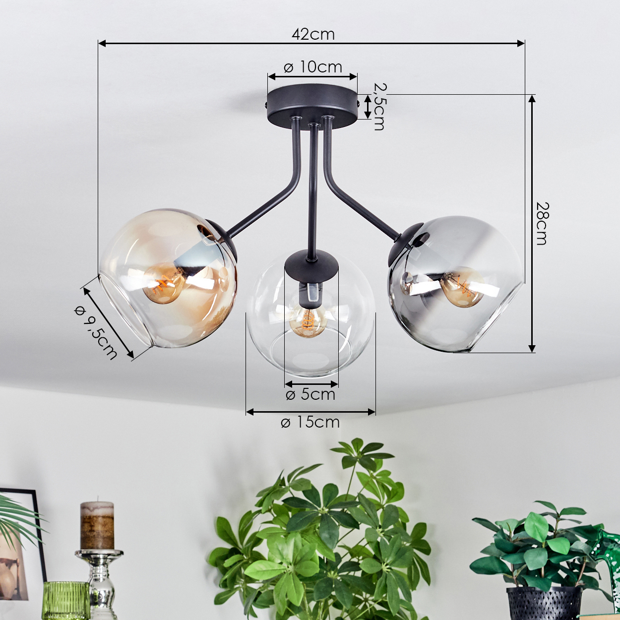 Eine moderne Deckenlampe mit drei Glaskugeln. Maße sind beschriftet. Pflanzen und Dekoration sind im Vordergrund sichtbar.