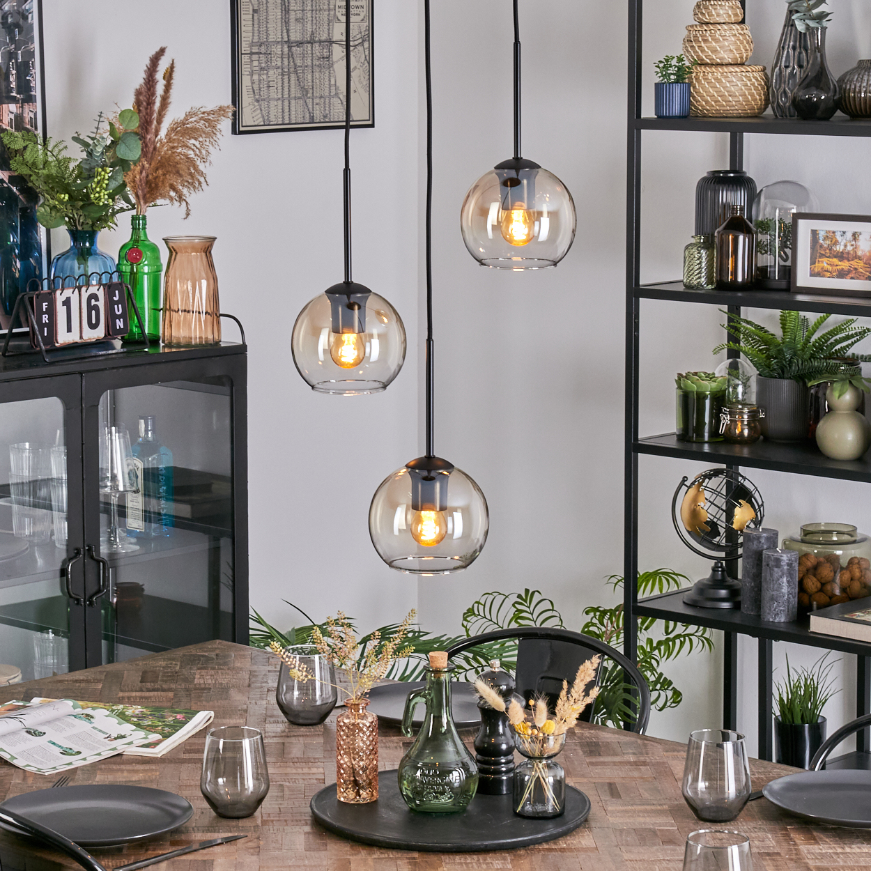 Esszimmer mit Hängelampen, Tisch mit Tellern, Gläsern und Dekoration. Regale mit Pflanzen und Dekorationsartikeln im Hintergrund.