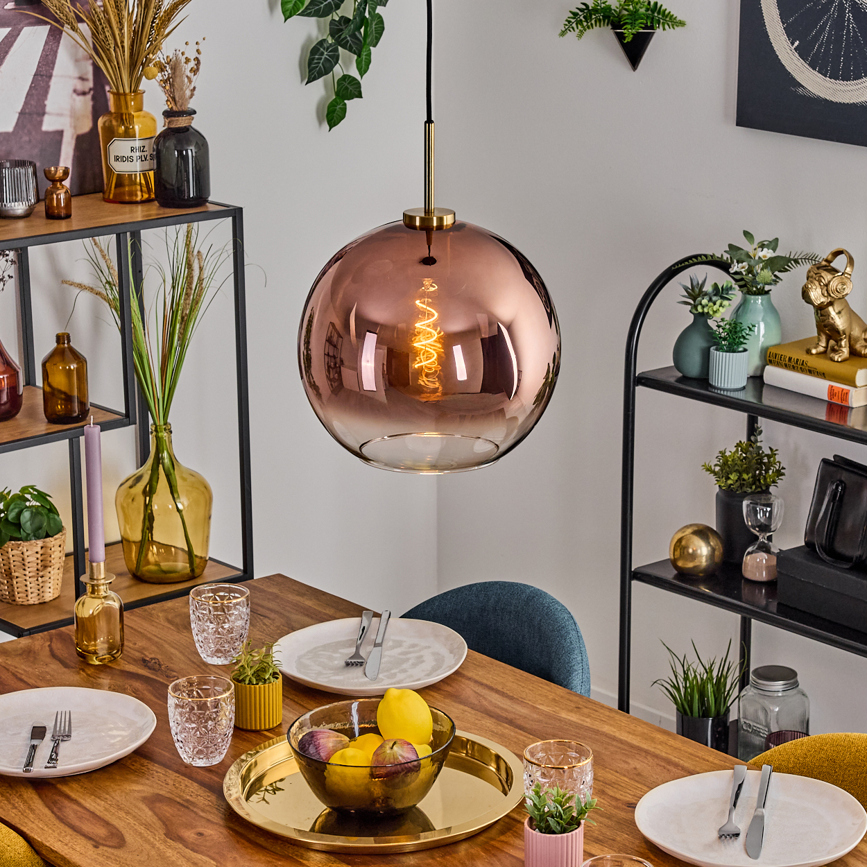 Ein Esstisch mit Tellern, Gläsern und einer Obstschale. Über dem Tisch hängt eine große, runde kupferfarbene Lampe.