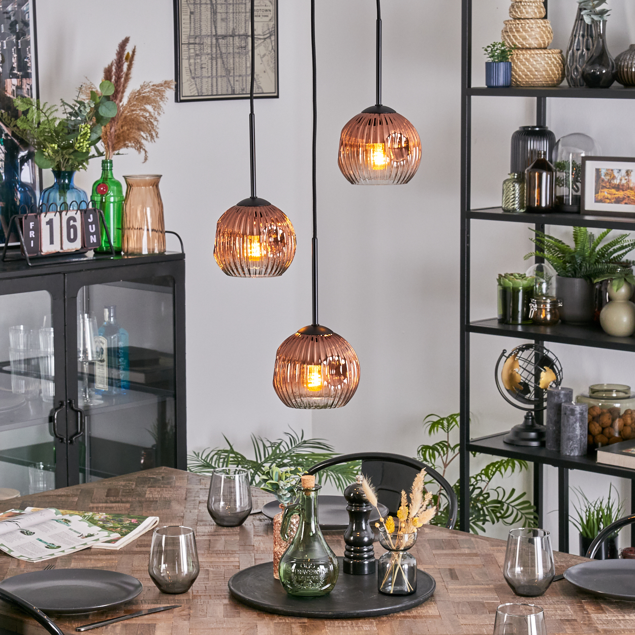 Esszimmer mit Holztisch, Glaslampen, Pflanzen und Regalen. Teller, Gläser und Dekorationsartikel sind auf dem Tisch arrangiert.