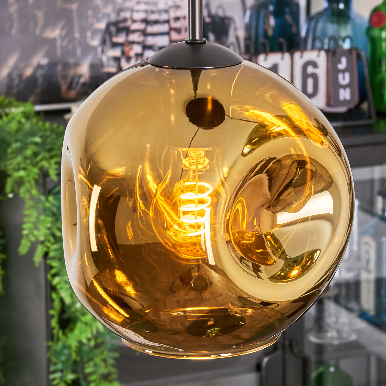 Eine goldene, blasenformige Pendelleuchte mit einer leuchtenden, spiralförmigen Glühbirne im Inneren. Im Hintergrund Pflanzen, Flaschen und ein Kalender.
