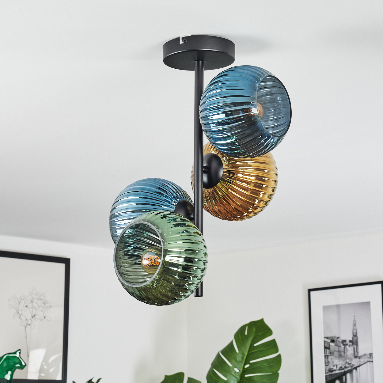 Eine moderne Deckenlampe mit vier bunten Glaskugeln: blau, gelb und grün. Sie hängt in einem weißen Raum mit Kunst und einer Pflanze.