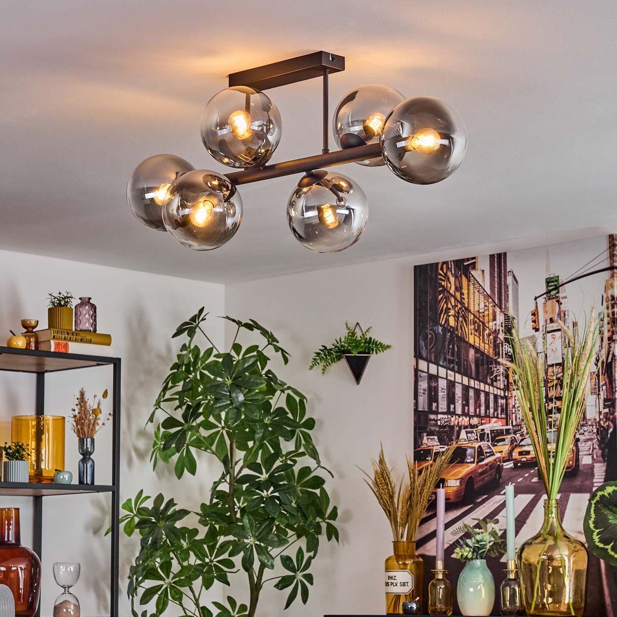 Ein modernes Wohnzimmer mit Kronleuchter, Pflanzen und einem Stadtbildgemälde. Auf einem Regal stehen verschiedene Vasen und Dekorationsartikel.