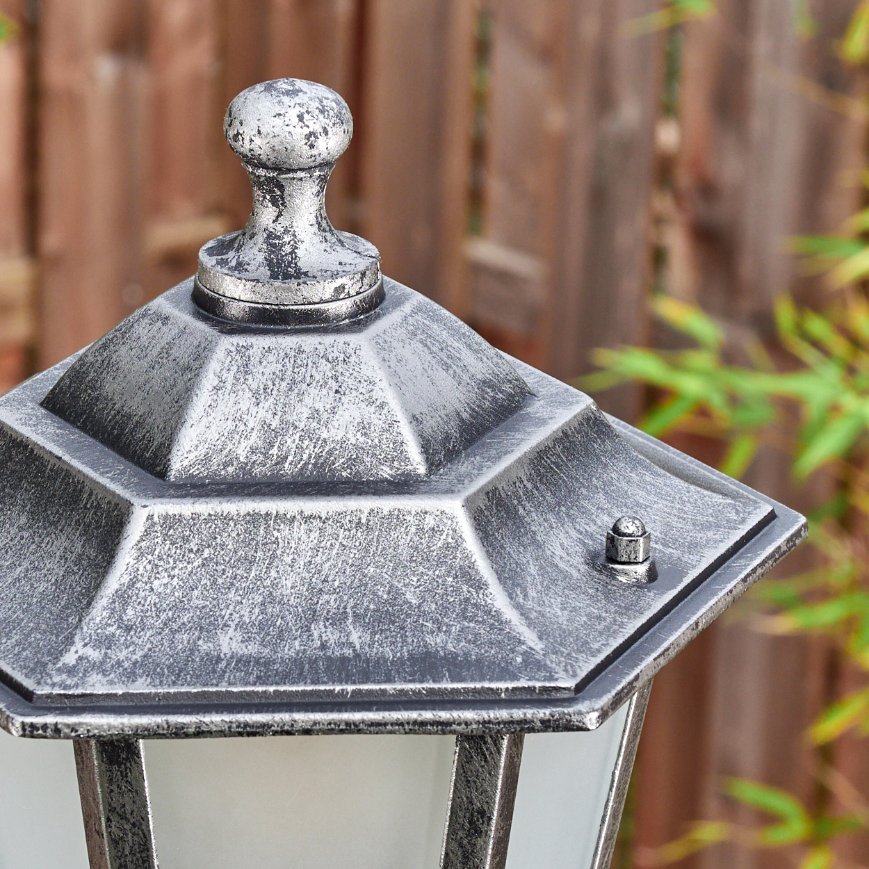 Nahaufnahme einer silbernen Außenlampe. Die Lampe hat eine geometrische Oberseite und einen kleinen Knopf. Im Hintergrund ist ein Holzzaun.
