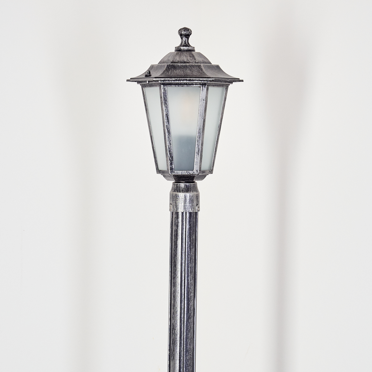 Eine dekorative silberne Straßenlaterne steht vor einem weißen Hintergrund. Die Lampe hat einen Schirm aus Milchglas und eine verzierte Spitze.