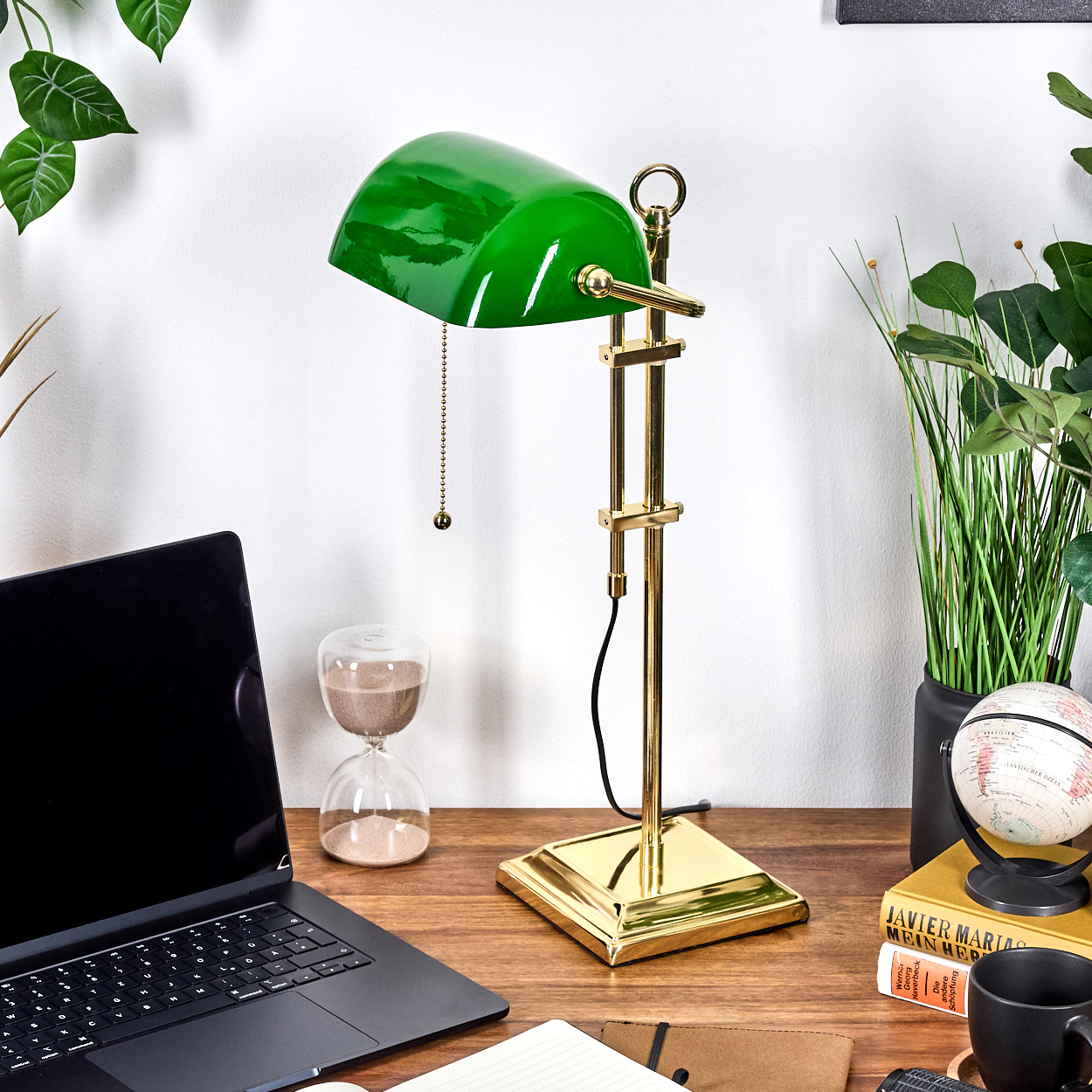 Eine grüne Bankerlampe steht auf einem Holzschreibtisch mit Laptop, Globus, Büchern und Pflanzen. Eine Sanduhr ist ebenfalls sichtbar.
