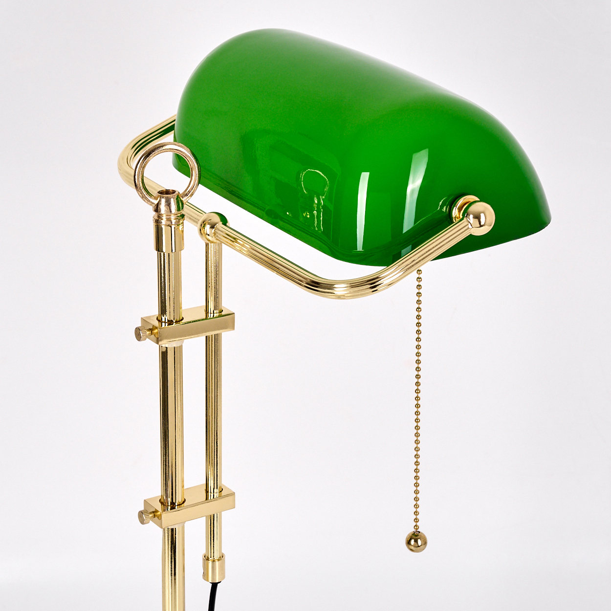 Eine grüne, glänzende Schreibtischlampe mit Messingrahmen und Zugkette, vor weißem Hintergrund.