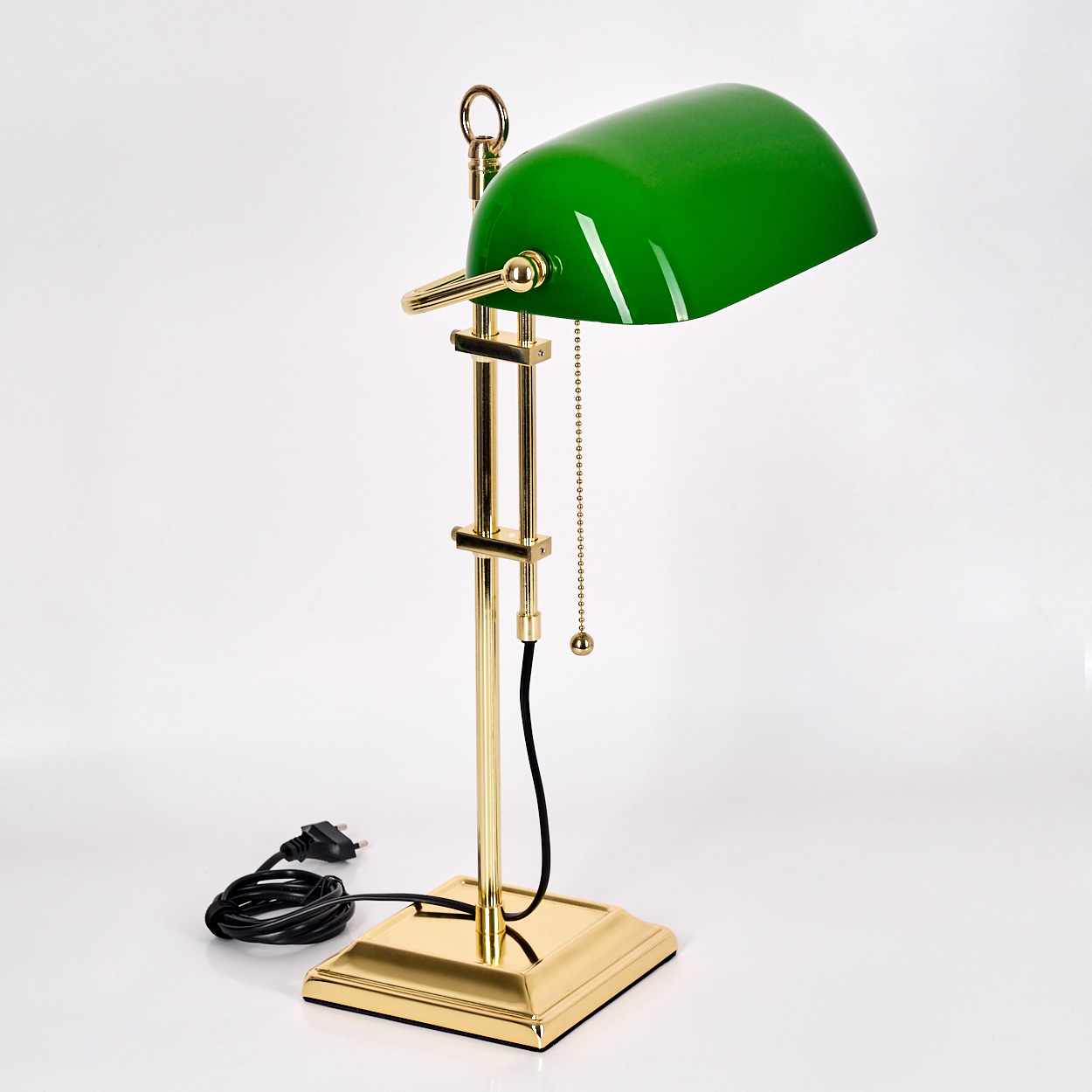 Eine grün schattierte, goldfarbene Bankerlampe steht auf einer weißen Oberfläche. Ein schwarzes Kabel und ein Stecker sind sichtbar.