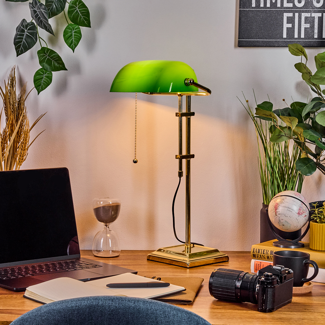 Ein Schreibtisch mit grüner Lampe, Laptop, Kamera, Büchern und einem Globus. Grüne Pflanzen im Hintergrund.