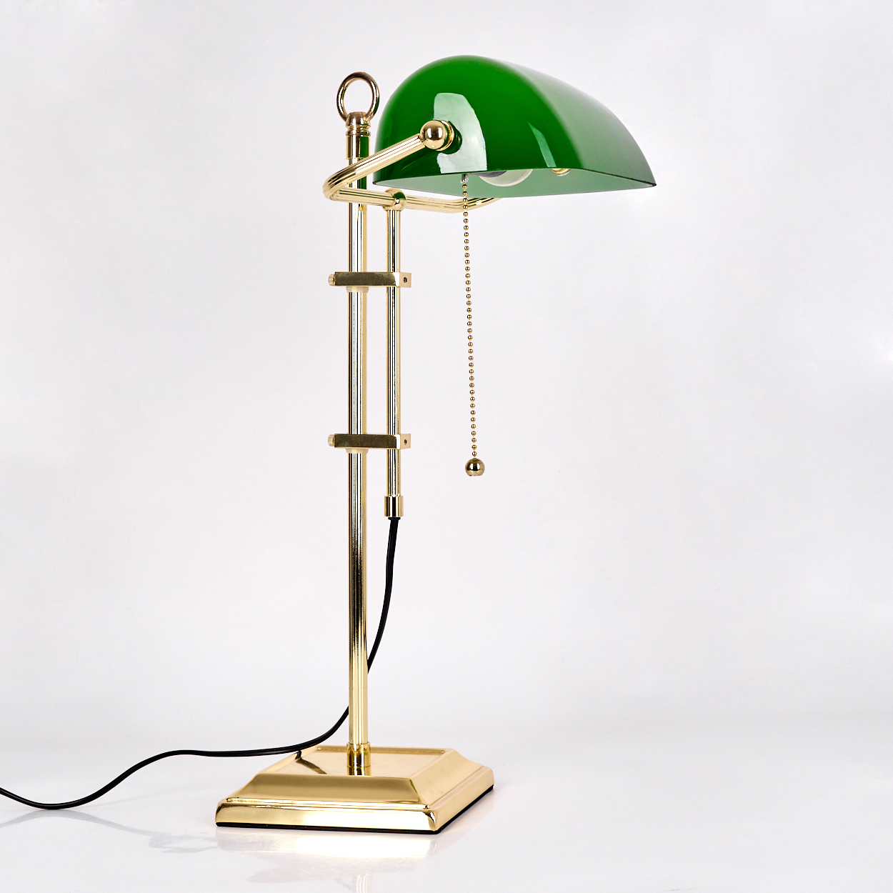 Eine grüne und goldene Schreibtischlampe. Die Lampe hat einen grünen Lampenschirm, einen goldenen Rahmen, eine Kette und einen quadratischen Goldsockel.