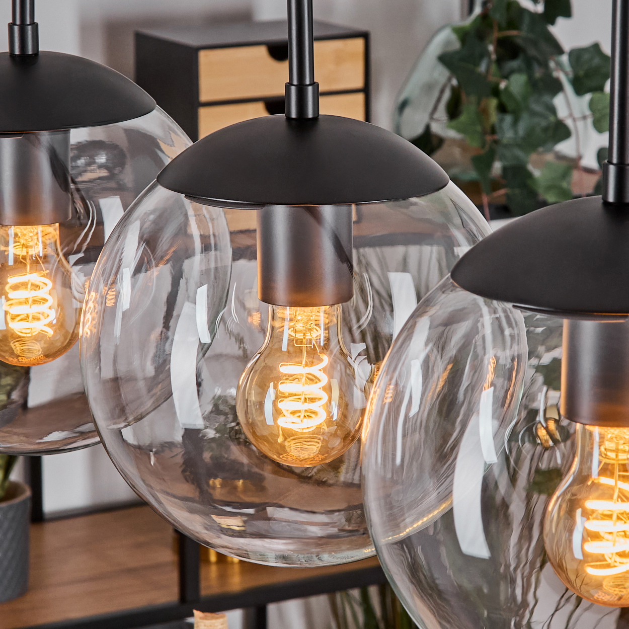 Drei Pendelleuchten mit Glaskugeln und Edison-Glühbirnen hängen. Schwarze Armaturen. Holzschrank und grüne Pflanze im Hintergrund.