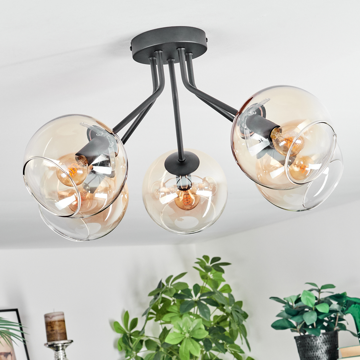 Eine moderne Deckenlampe mit drei bernsteinfarbenen Glaskugeln. Darunter stehen grüne Pflanzen an einer weißen Wand. Die Lampe hat schwarze Metallarme.