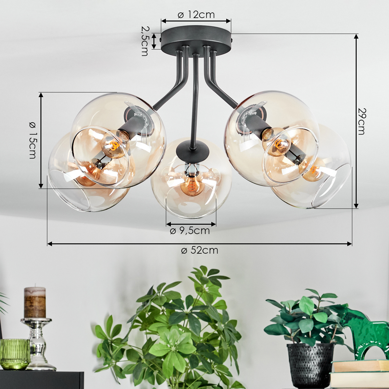 Moderne Deckenlampe mit fünf Glaskugeln, Maße beschriftet. Unten sind grüne Pflanzen und ein Kerzenhalter zu sehen.