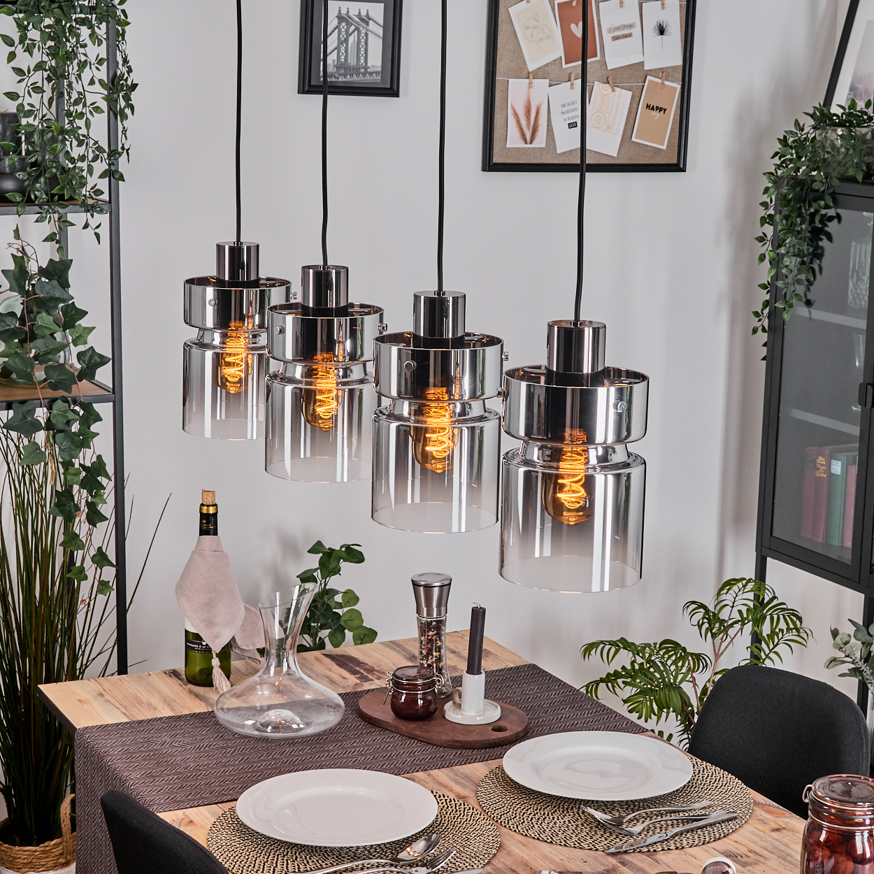 Esszimmer mit vier Pendelleuchten über einem Holztisch, der zum Abendessen gedeckt ist. Pflanzen, Wein und Dekorationen sind sichtbar.