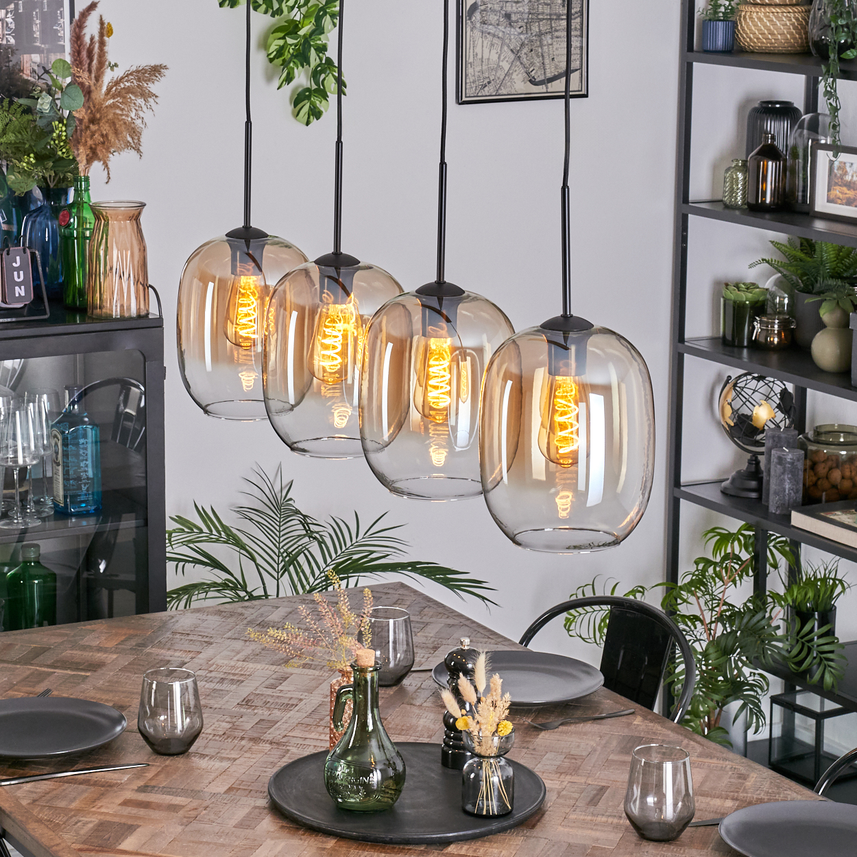 Esszimmer mit Holztisch, Hängelampen, Pflanzen und Regalen mit Dekorationen. Teller, Gläser und Blumen stehen auf dem Tisch.