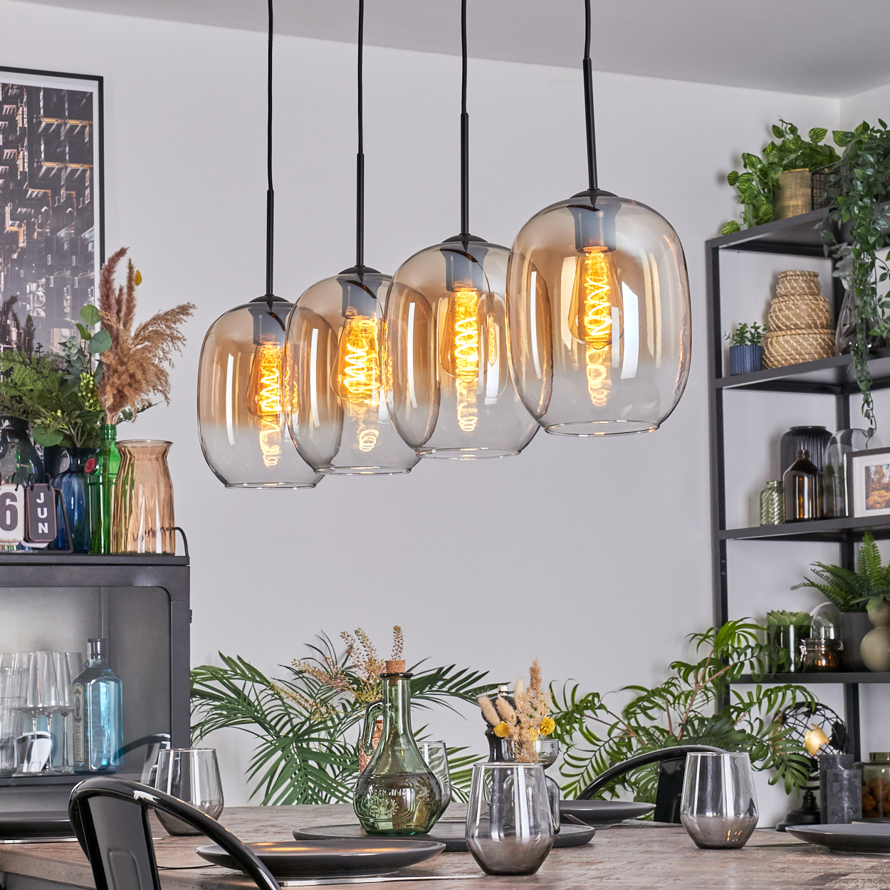Vier Pendelleuchten aus bernsteinfarbenem Glas hängen über einem Holztisch, der zum Abendessen gedeckt ist. Pflanzen und Regale sind im Hintergrund zu sehen.