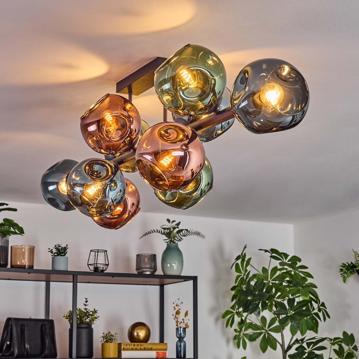 Eine moderne Deckenleuchte mit mehreren Glaskugeln in Grün-, Kupfer- und Grautönen. Sie hängt an einer weißen Decke, darunter ein Regal.