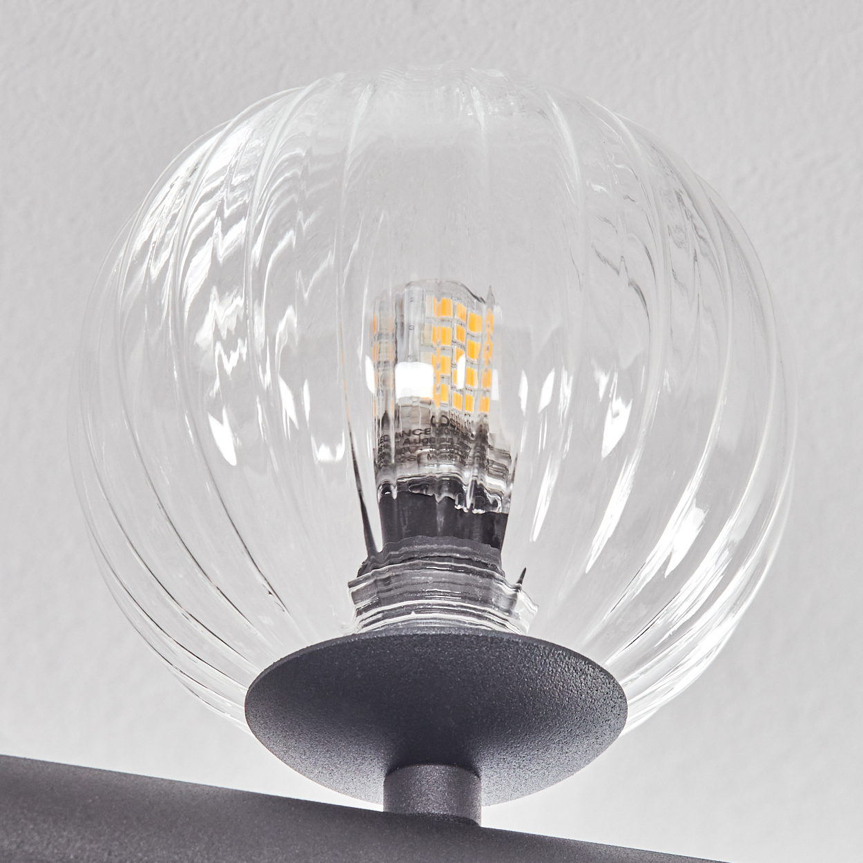 Eine klare, gerippte Glaskugel-Leuchte. Im Inneren ist eine leuchtende LED-Glühbirne sichtbar. Die Leuchte ist auf einem grauen Metallsockel montiert.