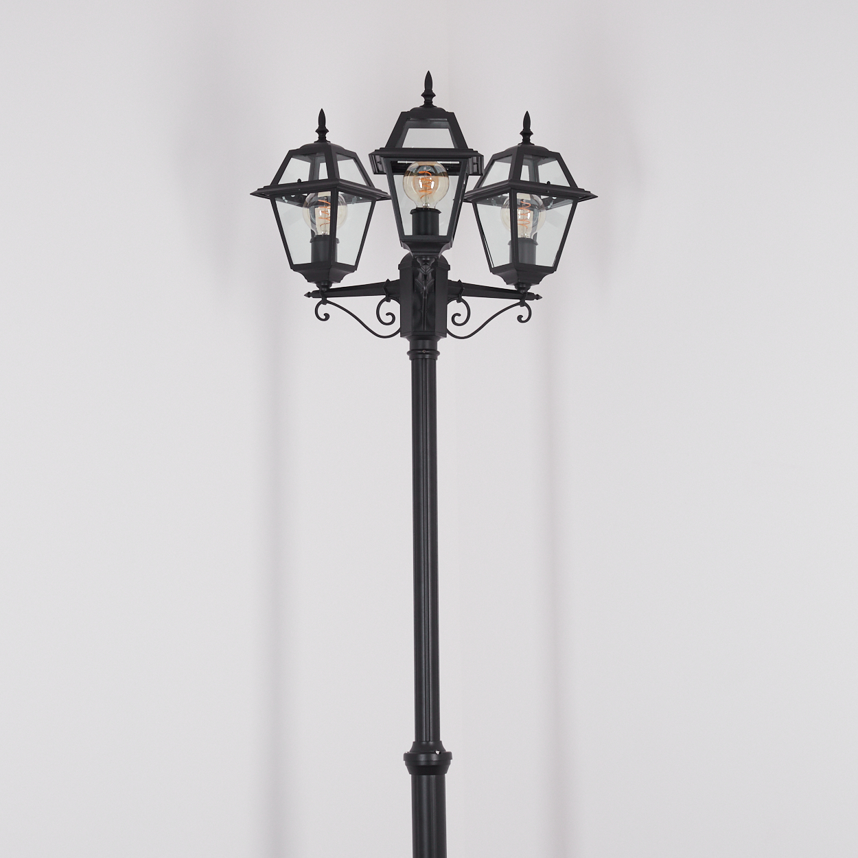 Eine schwarze Straßenlaterne mit drei Glaskästen. Sie hat eine hohe Stange und verzierte Details auf weißem Hintergrund.