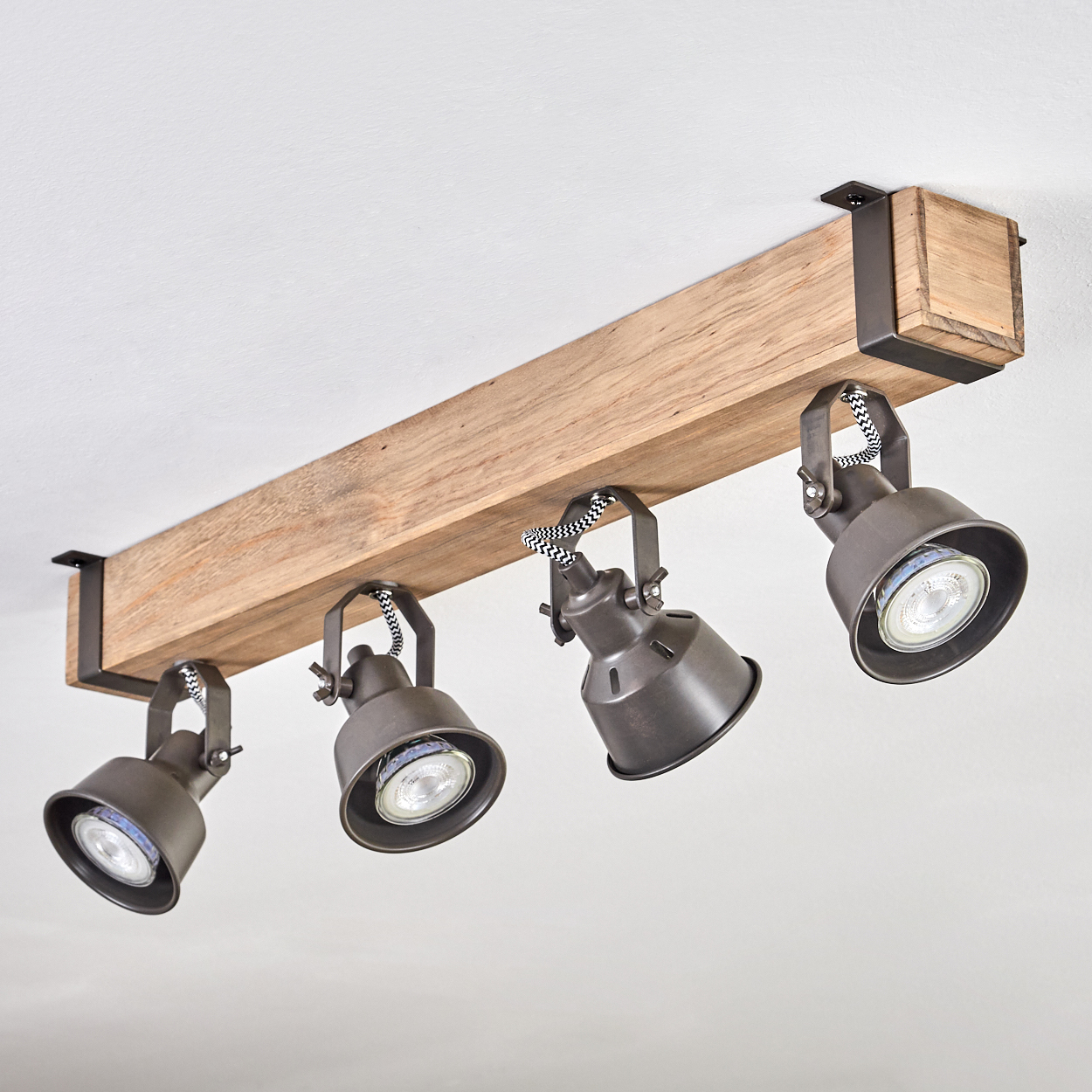 Ein Holzbalken mit vier grauen Strahlern. Die Lampen haben schwarz-weiß gestreifte Kabel und sind an einer weißen Decke befestigt.