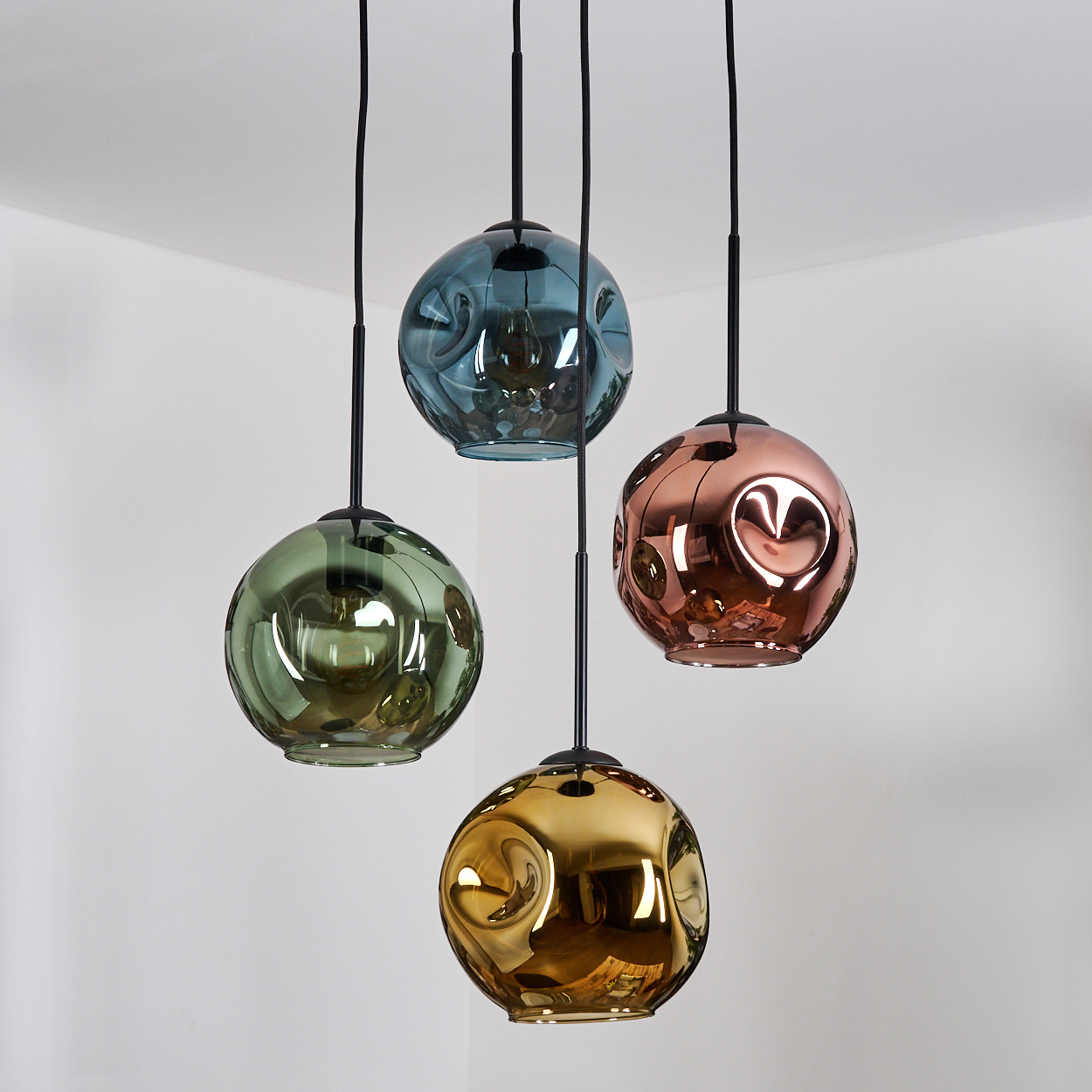 Vier bunte, kugelförmige Pendelleuchten hängen an schwarzen Kabeln. Jede hat eine einzigartige Farbe: grün, blau, kupfer und gold. Der Hintergrund ist weiß.
