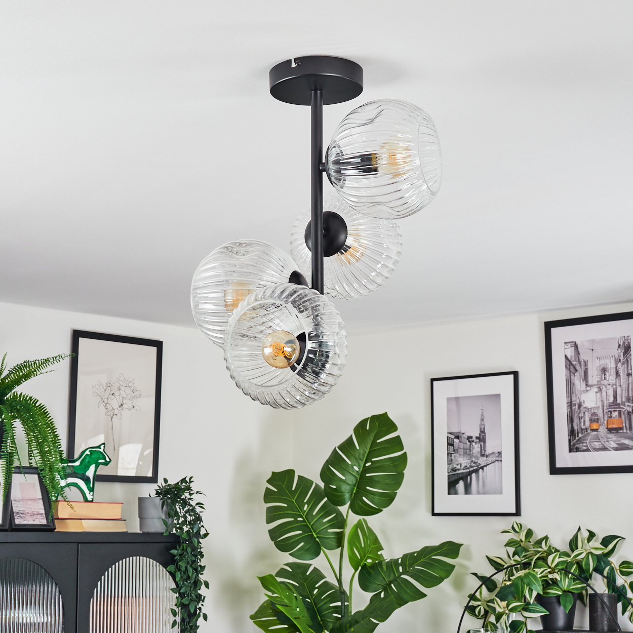 Eine moderne Hängelampe mit vier Glaskugelschirmen hängt von der Decke. Dekorative Pflanzen und gerahmte Bilder im Hintergrund.