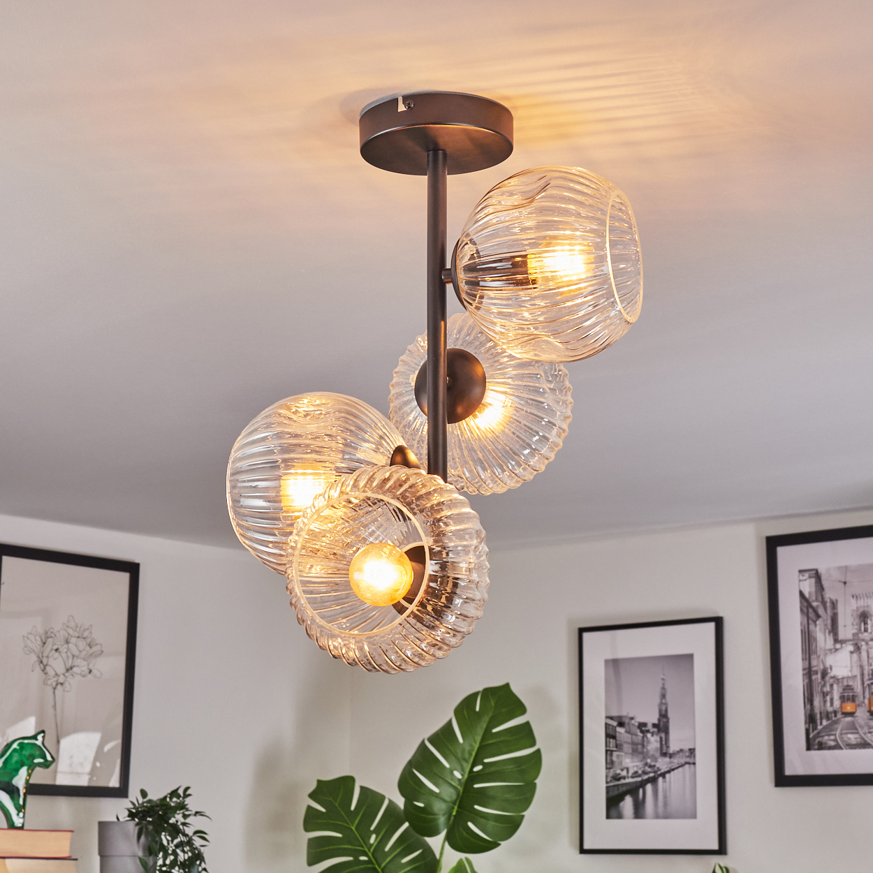 Eine moderne Deckenlampe mit vier Glasschirmen. Die Schirme haben ein geripptes Design. Der Hintergrund hat gerahmte Bilder und Pflanzen.