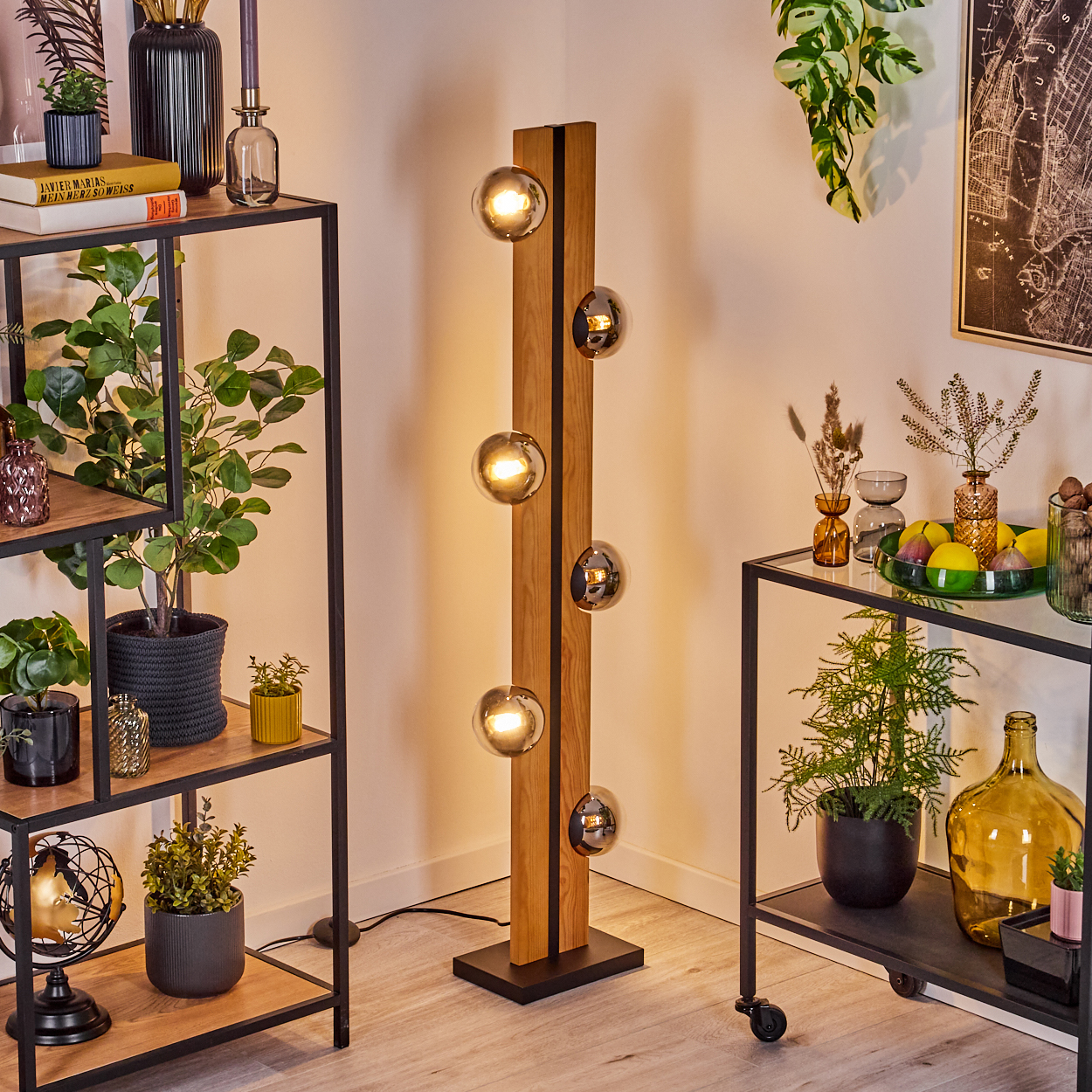 Ein modernes Zimmer mit einer Holzlampe mit Glaskugeln, Pflanzen und Regalen voller Dekoration. Rechts ein Wagen mit Pflanzen.