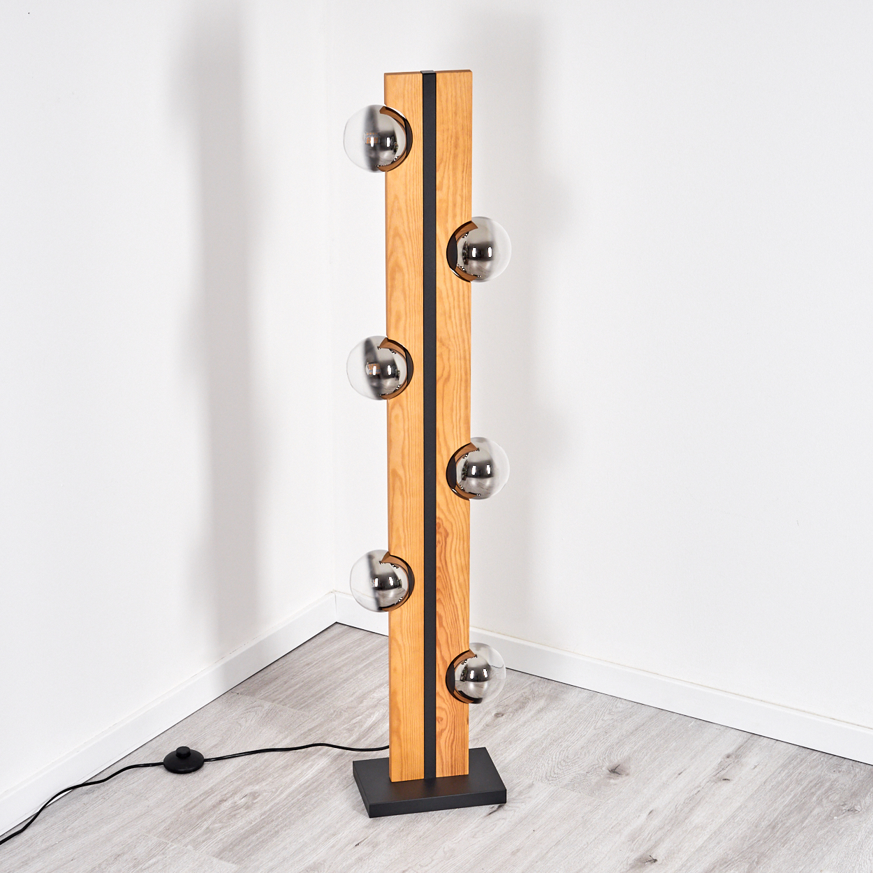 Eine moderne Stehlampe mit Holzrahmen und sechs kugelförmigen Glühbirnen. Sie steht auf einem schwarzen Sockel vor einer weißen Wand.
