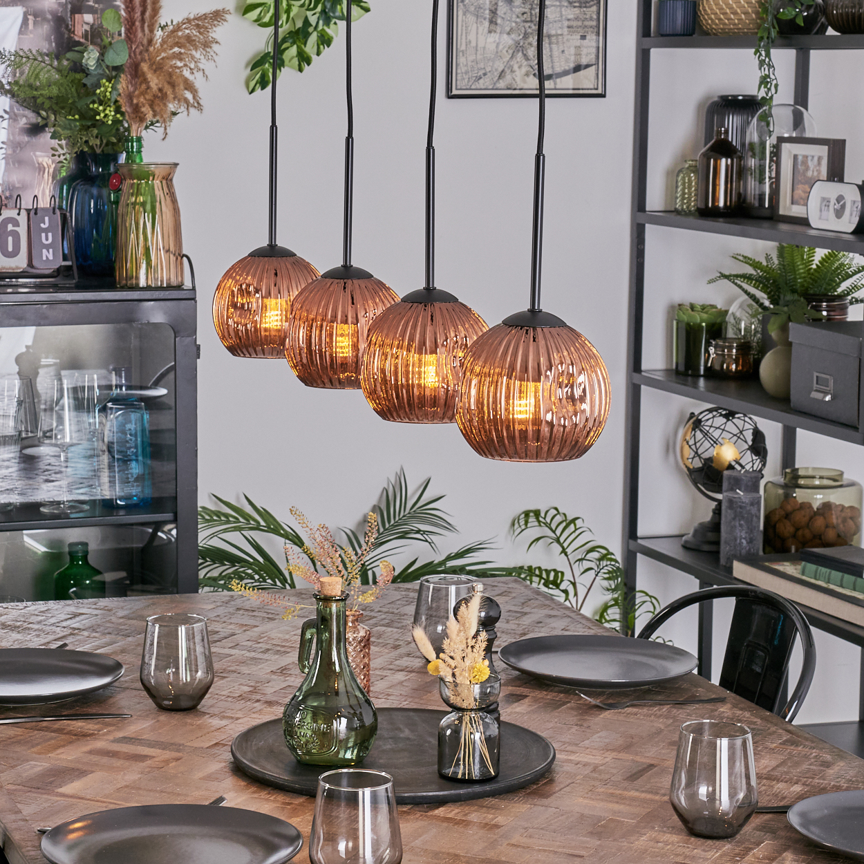 Esszimmer mit Holztisch, Kupferlampen und Dekorationen. Teller und Gläser sind gedeckt. Regale mit Pflanzen im Hintergrund.