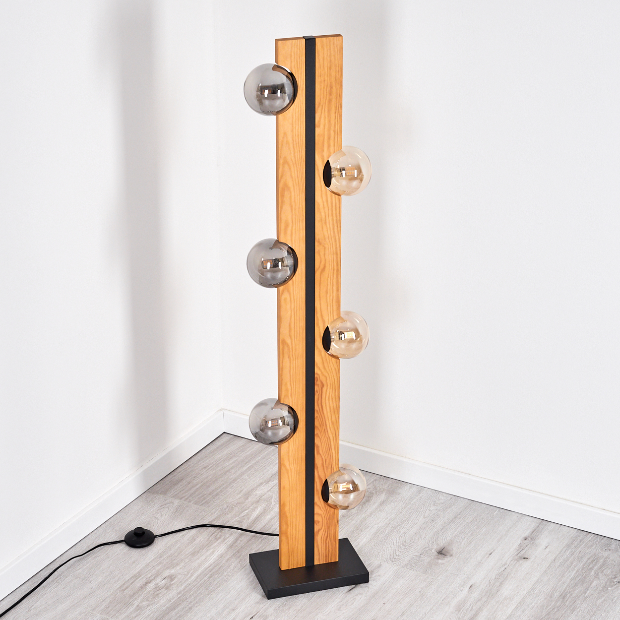 Eine moderne Stehlampe mit Holzbalken, schwarzen Akzenten und Glaskugellampen. Sie steht auf einem schwarzen Sockel.