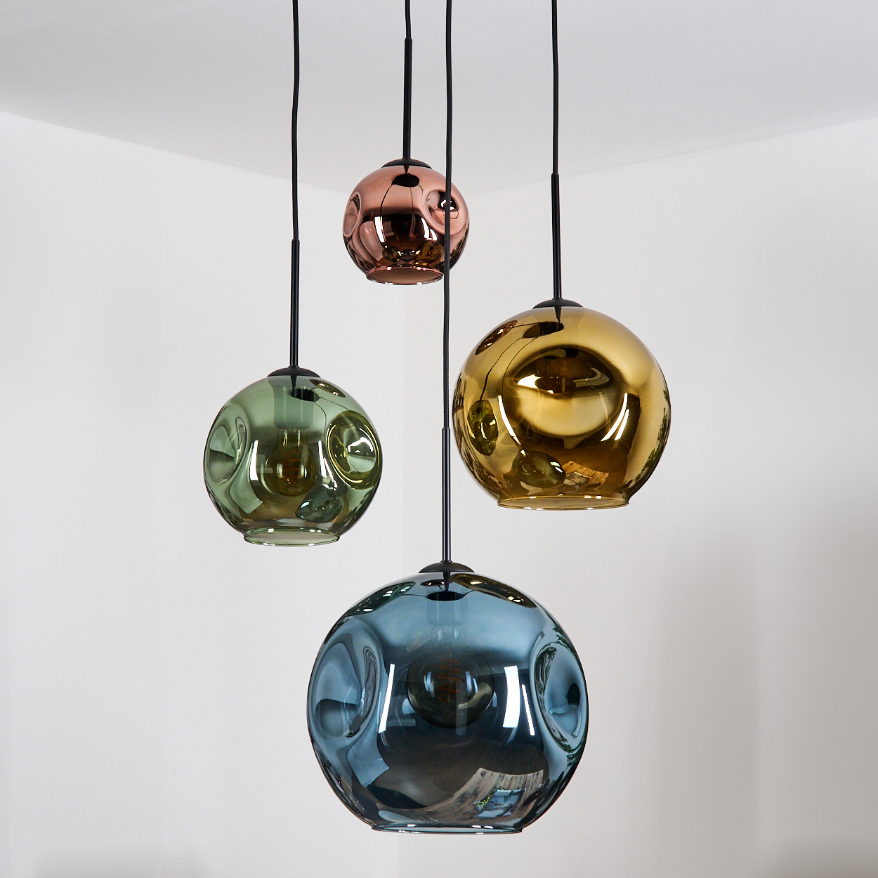 Vier kugelförmige Pendelleuchten in Grün, Gold, Kupfer und Blau hängen von der Decke vor einer weißen Wand.