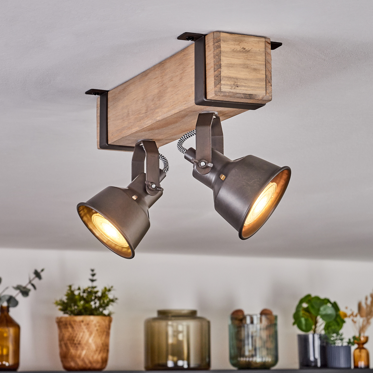 Deckenstrahler mit zwei Lampen, Holzbalken und schwarzen Halterungen. Pflanzen und Dekor darunter.