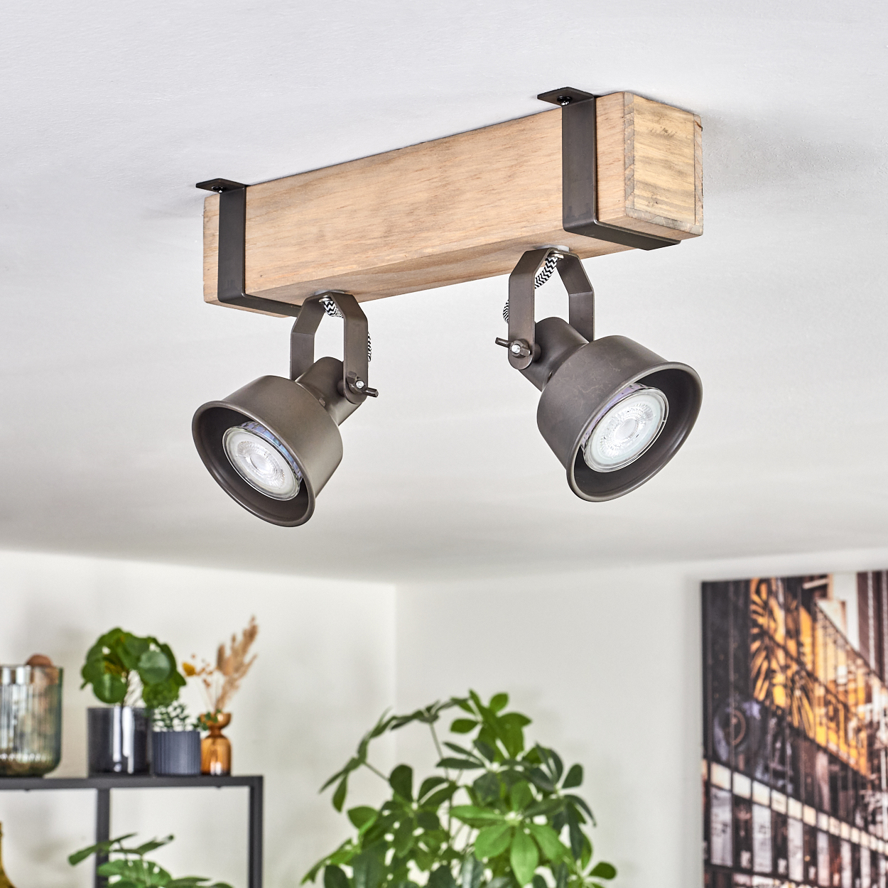 An der Decke montierte Leuchte mit zwei Strahlern, Holzbalken und Metallhalterungen. Pflanzen und Kunstwerke im unscharfen Hintergrund.