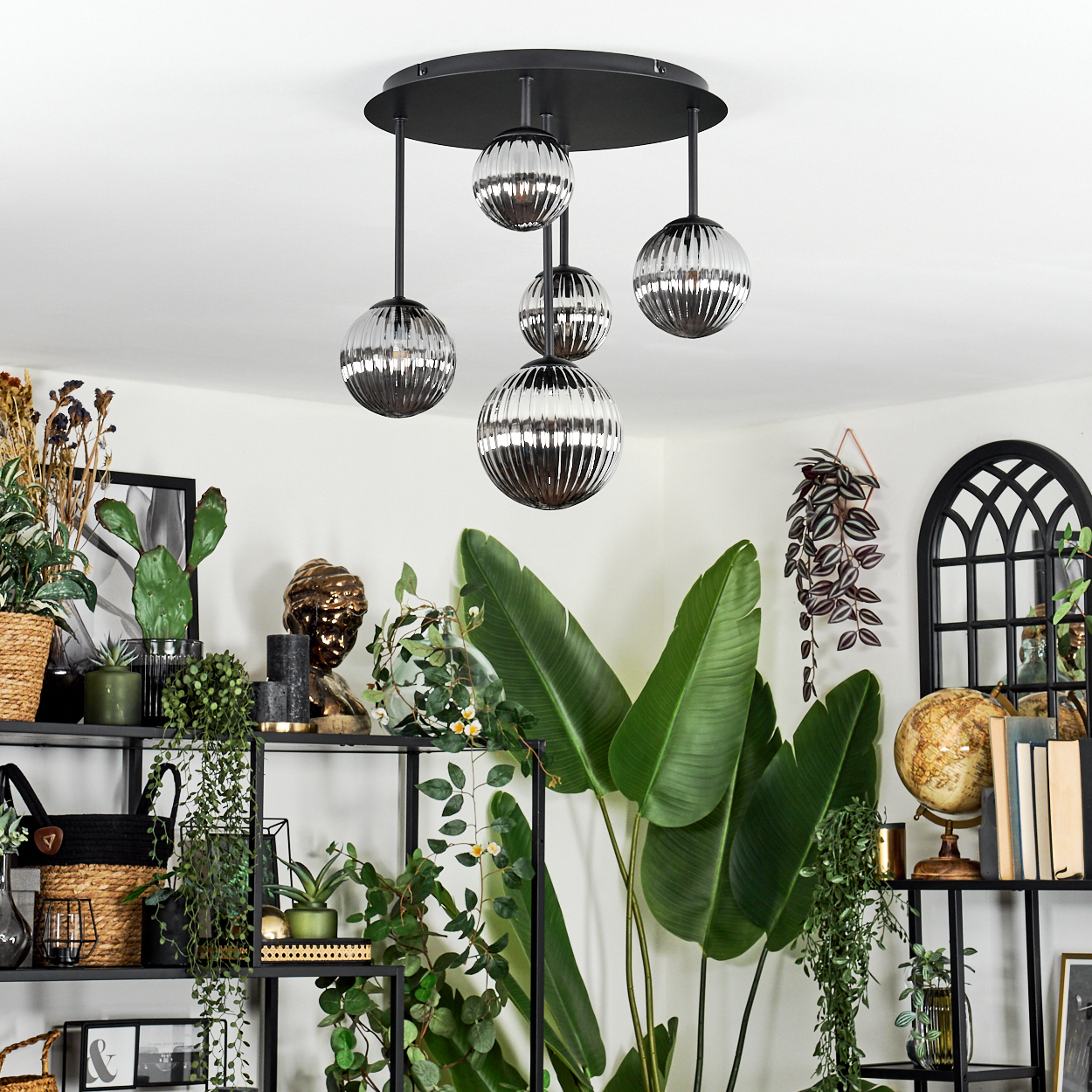 Ein modernes Wohnzimmer mit einem schwarzen Metallregal voller Pflanzen und Dekoration. Eine Leuchte hängt von der Decke.