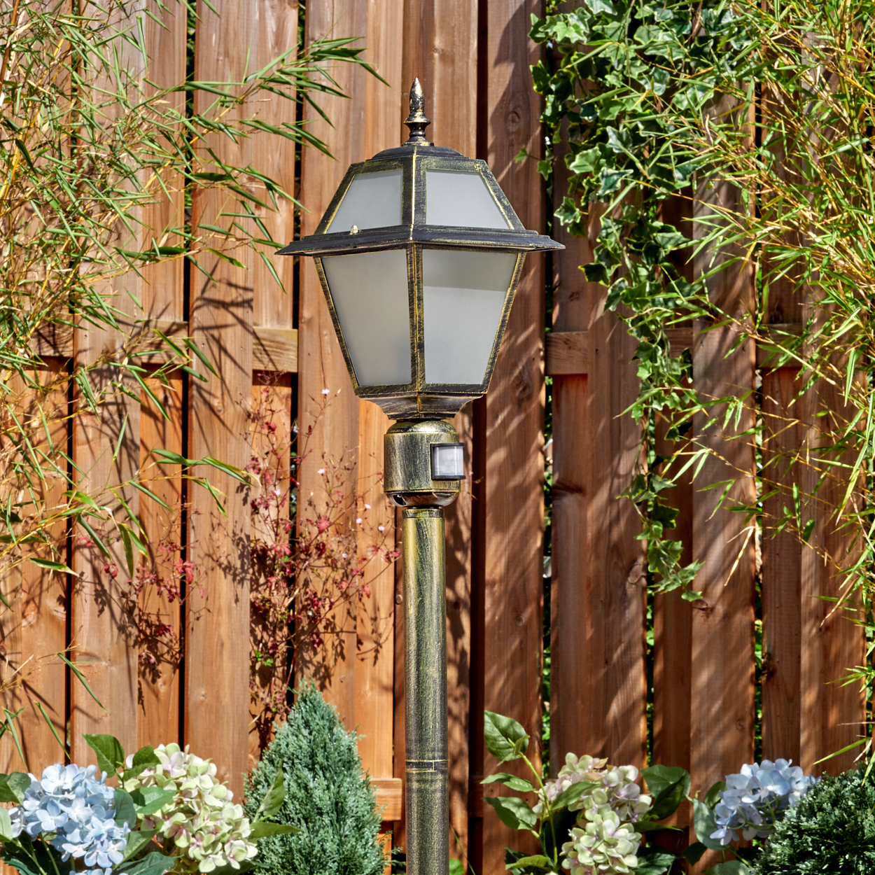 Eine dekorative Außenlampe steht vor einem Holzzaun. Pflanzen und Blumen umgeben die Lampe. Bambus und Efeu sind ebenfalls vorhanden.