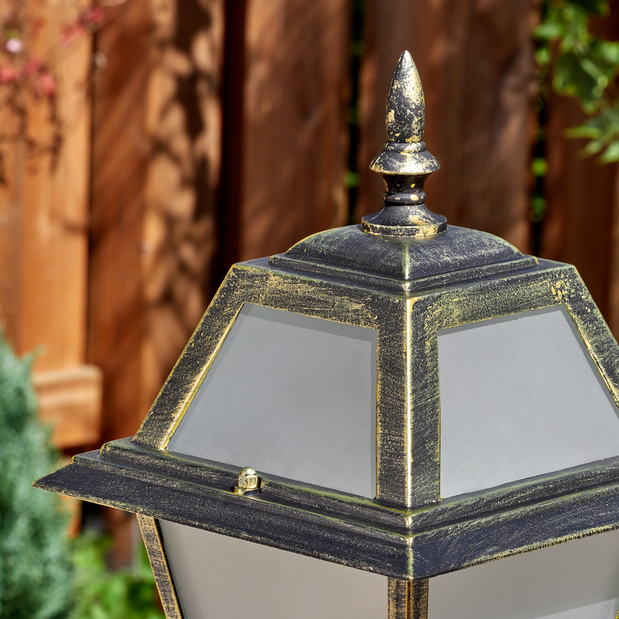 Nahaufnahme einer dekorativen Außenlampe mit schwarzen und goldenen Akzenten. Im Hintergrund ein Holzzaun und Grün.