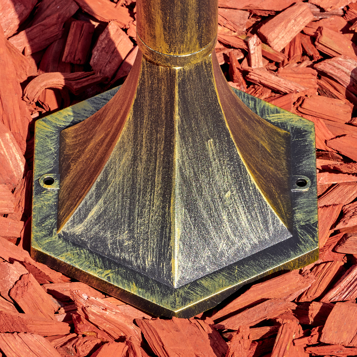 Ein dekorativer Lampensockel in Gold und Schwarz liegt auf roter Mulch. Die Basis hat eine sechseckige Form mit konischer Spitze.