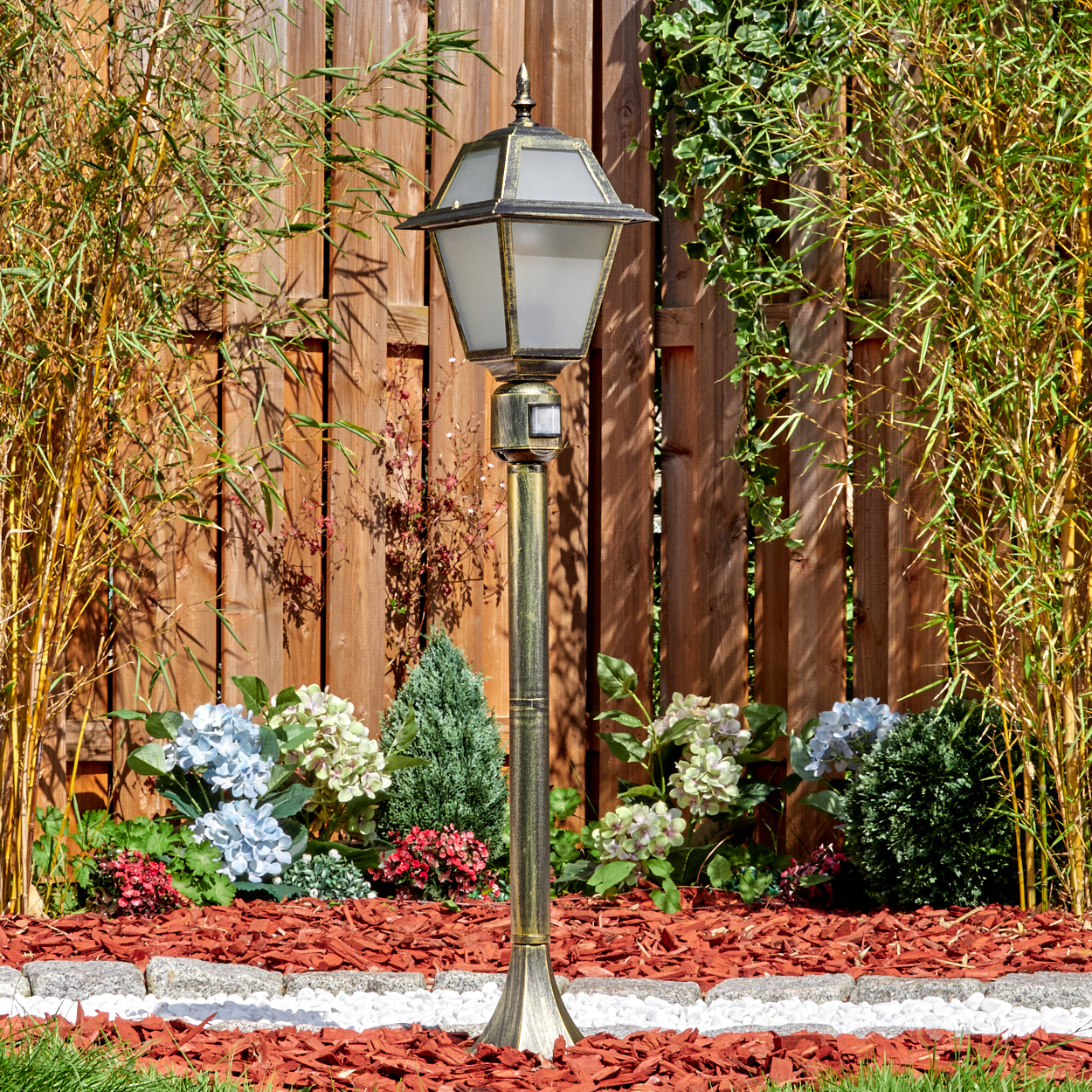 Eine dekorative Außenlampe steht in einem Garten mit Blumen, Bambus und einem Holzzaun im Hintergrund.