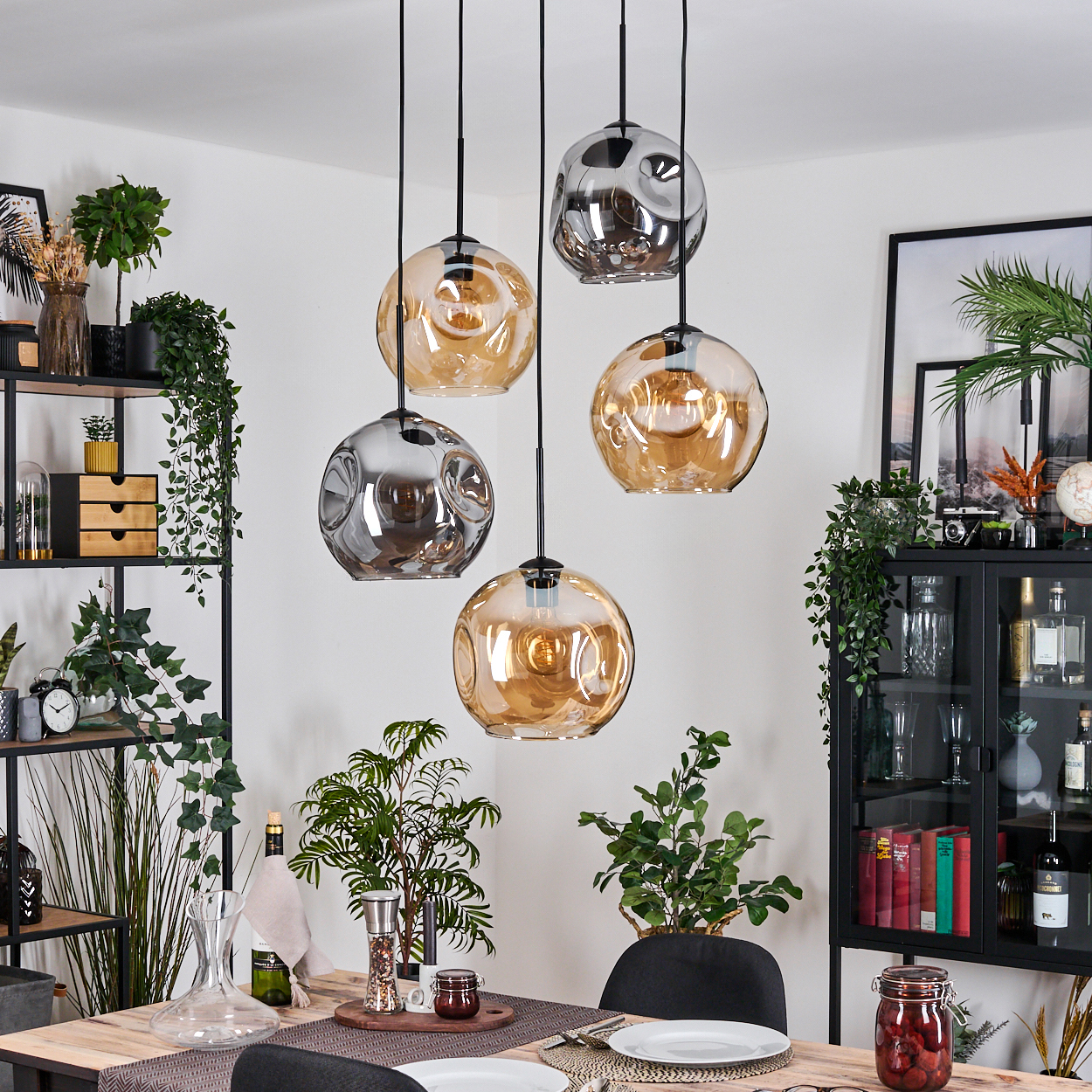 Esszimmer mit Hängelampen. Ein Tisch ist mit Tellern und Dekoration gedeckt. Regale mit Pflanzen und ein Schrank mit Gegenständen sind sichtbar.