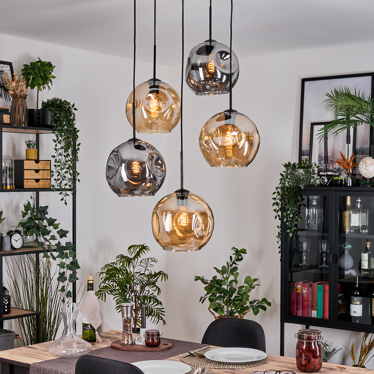 Esszimmer mit Hänge-Kugelleuchten, Pflanzen, Tischgedeck und schwarzem Regal. Die Lampen haben freiliegende Glühbirnen.