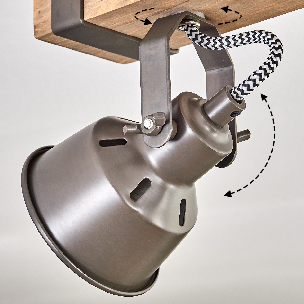 Ein Metall-Spotlight ist mit einem gemusterten Kabel an einem Holzbalken befestigt. Der Scheinwerfer ist nach unten geneigt. Pfeile zeigen die Drehung.