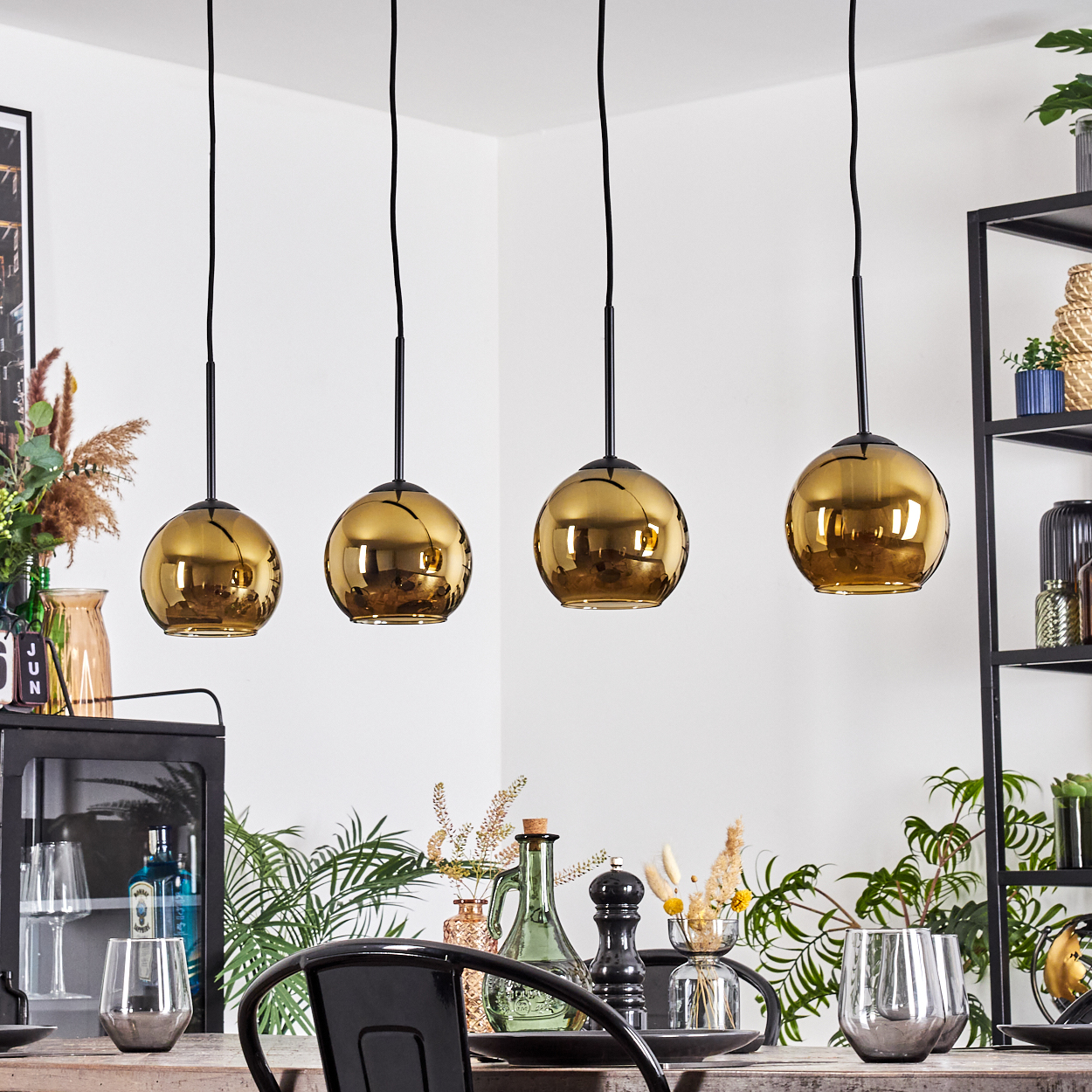 Vier goldene Pendelleuchten hängen über einem Tisch. Die Szene umfasst Pflanzen, Gläser und ein schwarzes Metallregal.