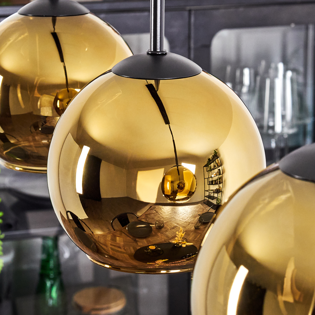 Drei goldene, kugelförmige Pendelleuchten hängen. In den Kugeln spiegeln sich ein Raum mit Holzböden, Möbeln und Regalen.