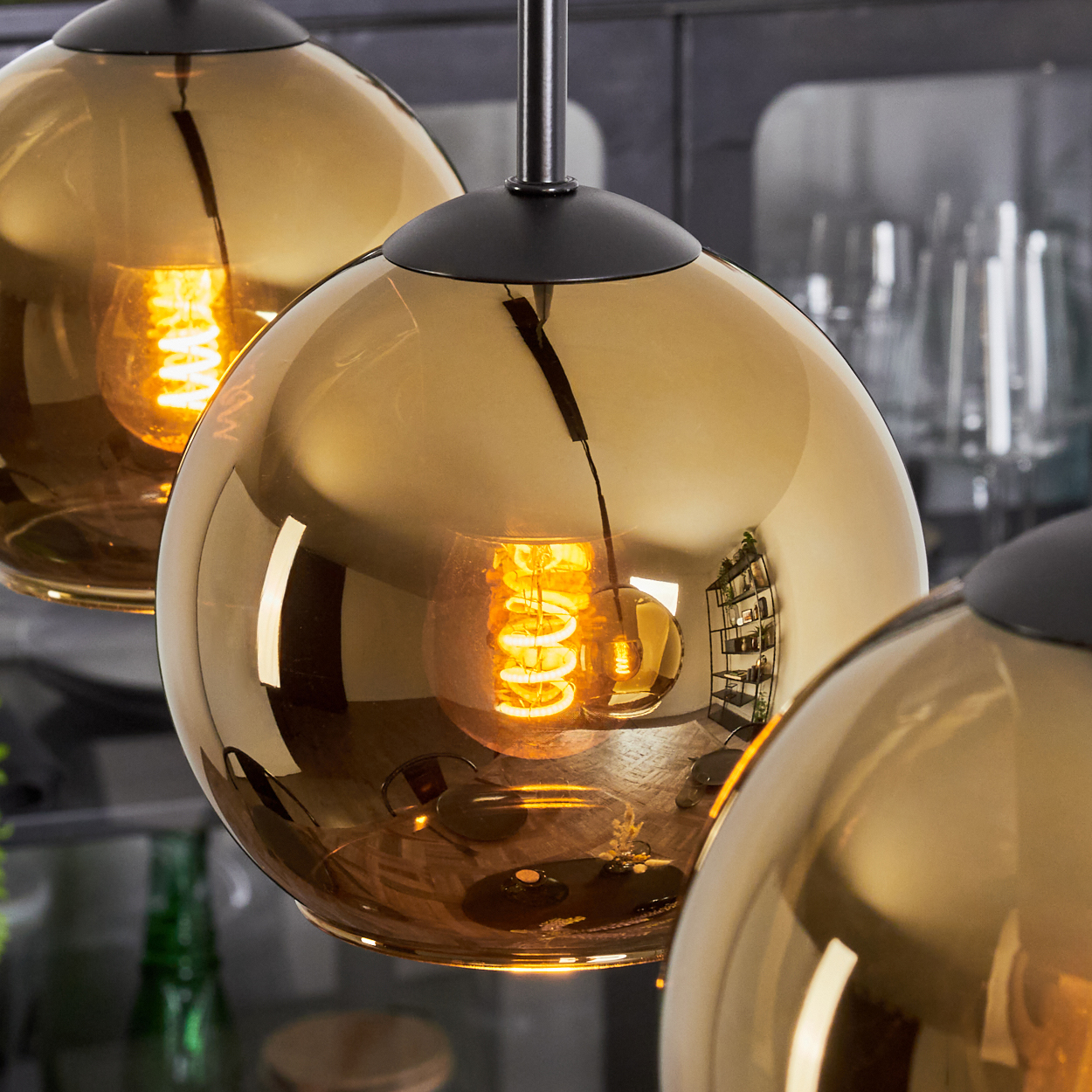 Drei goldene kugelförmige Pendelleuchten mit sichtbaren Glühbirnen. Schwarze Armaturen. Spiegelung von Regalen und Gläsern im Hintergrund.