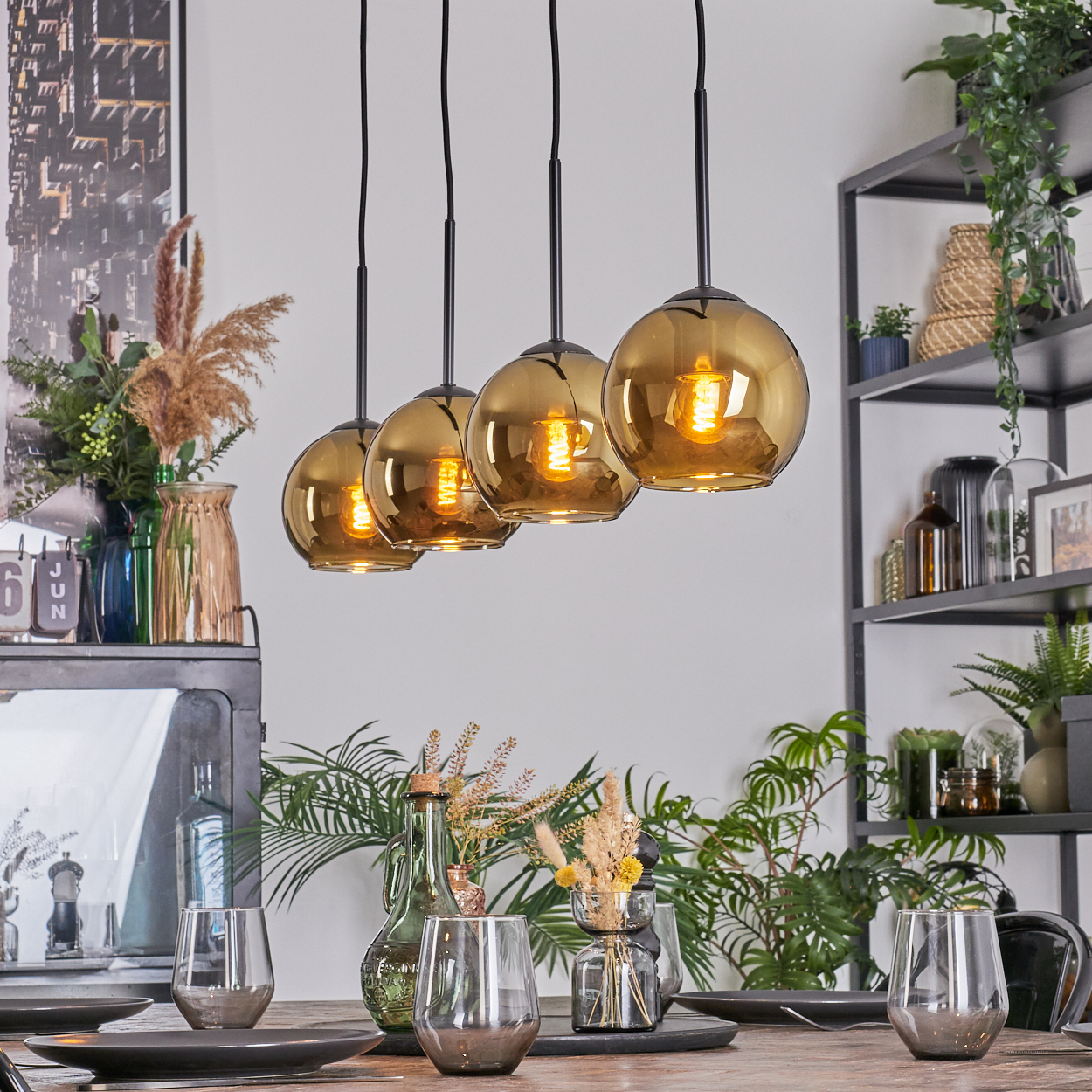 Esszimmer mit hängenden, bernsteinfarbenen Glasleuchten. Tisch mit Gläsern und Tellern gedeckt. Regale mit Pflanzen im Hintergrund.