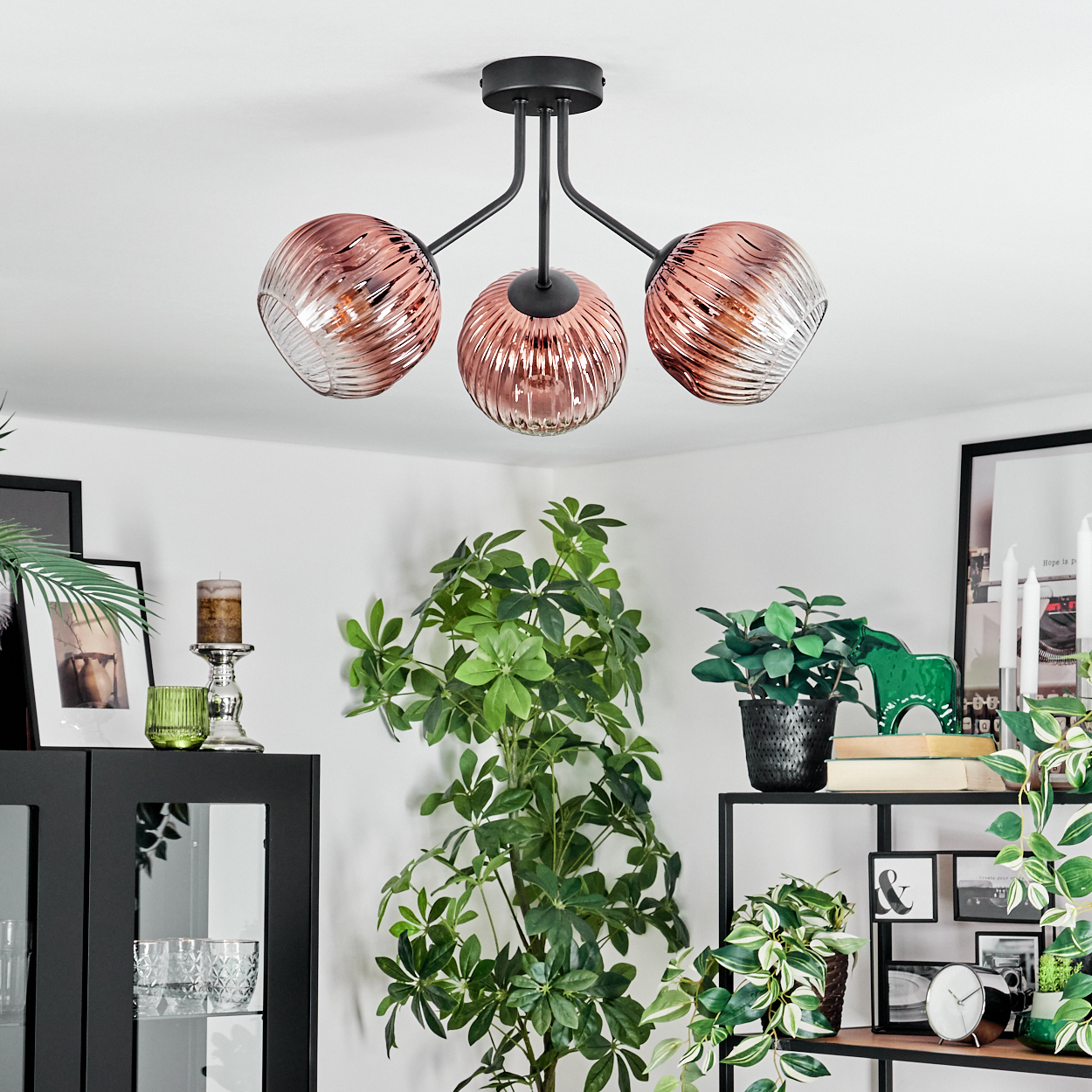 Eine moderne Deckenlampe mit drei rosa Glaskugeln hängt über einem Raum mit Pflanzen und Dekorationen. Schwarze Möbel im Hintergrund.