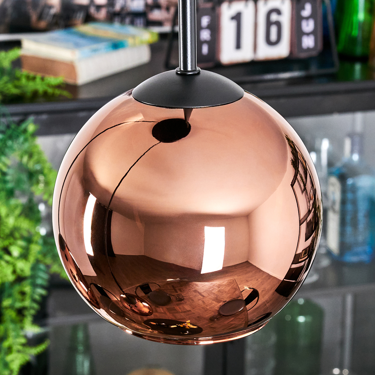 Eine kupferfarbene, kugelförmige Lampe hängt. Sie reflektiert den Raum und einen Kalender mit dem 16. Grüne Pflanzen und Flaschen im Hintergrund.